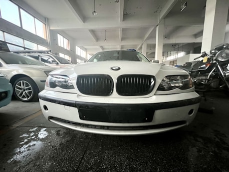 A white car is parked indoors in what appears to be a garage or showroom. The setting includes other parked cars and a motorcycle visible in the background. The environment is well-lit with large windows, and the floor shows some reflections from water or oil. The car's front view is prominently displayed with emphasis on its grille and headlights.