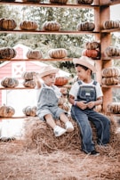 Two children wearing straw hats and denim overalls sit on a hay bale surrounded by an array of pumpkins. The setting is rustic and outdoors, with a wooden structure displaying the pumpkins in neat rows. A red barn-like building is visible in the background amid lush greenery.