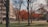 A campus scene with a large building on the left. Several trees, some with autumnal orange leaves, are scattered across a lawn. Paths and walkways run through the area, with a few people visible walking or standing around. There are also several benches and a lamppost near the building.