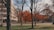 A campus scene with a large building on the left. Several trees, some with autumnal orange leaves, are scattered across a lawn. Paths and walkways run through the area, with a few people visible walking or standing around. There are also several benches and a lamppost near the building.
