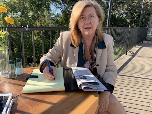 A person is seated at a wooden table outdoors, writing in a notebook with a pen. They are wearing glasses and a light-colored blazer with a scarf. A newspaper is partially visible on the table, and there is a vase with yellow flowers and salt and pepper shakers nearby. The background features a metal railing and lush green trees, suggesting a park or garden setting.