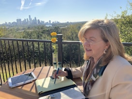 A woman is sitting at an outdoor table overlooking a city skyline. She appears engaged and thoughtful, writing in a notebook. Bright yellow flowers in a vase and an electronic tablet are on the table. The setting suggests a tranquil environment, with greenery in the background and a clear sky.