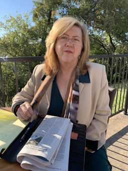 A person with light-colored hair wearing glasses and a beige blazer sits outdoors by a table with papers and a pen on it. The background features green trees and a railing, suggesting a park or garden setting.