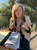 An older woman with blonde hair wearing glasses sits at an outdoor table. She holds a scarf in her hands and wears a light-colored blazer over a dark top. A newspaper, notebook, and pen are on the table, with a metal railing and greenery in the background.
