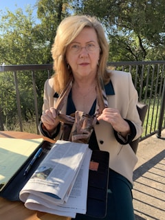 An older woman with blonde hair wearing glasses sits at an outdoor table. She holds a scarf in her hands and wears a light-colored blazer over a dark top. A newspaper, notebook, and pen are on the table, with a metal railing and greenery in the background.