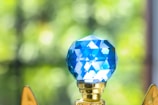 A close-up view of a blue, faceted crystal object mounted on a golden base. The crystal reflects light, creating a geometric pattern. In the background, soft-focus greenery is visible, providing a vibrant contrast.
