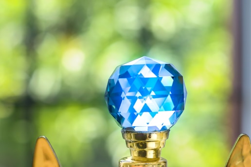 A close-up view of a blue, faceted crystal object mounted on a golden base. The crystal reflects light, creating a geometric pattern. In the background, soft-focus greenery is visible, providing a vibrant contrast.