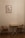 A small, minimalist interior scene features a wooden table with two matching wooden chairs with brown cushions on either side. A framed artwork of a Moka pot hangs on the beige wall above the table, adding a touch of vintage charm. The wooden floor complements the warm, neutral tones of the furniture and wall.