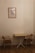A small, minimalist interior scene features a wooden table with two matching wooden chairs with brown cushions on either side. A framed artwork of a Moka pot hangs on the beige wall above the table, adding a touch of vintage charm. The wooden floor complements the warm, neutral tones of the furniture and wall.