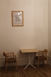 A small, minimalist interior scene features a wooden table with two matching wooden chairs with brown cushions on either side. A framed artwork of a Moka pot hangs on the beige wall above the table, adding a touch of vintage charm. The wooden floor complements the warm, neutral tones of the furniture and wall.