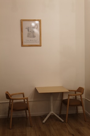 A small, minimalist interior scene features a wooden table with two matching wooden chairs with brown cushions on either side. A framed artwork of a Moka pot hangs on the beige wall above the table, adding a touch of vintage charm. The wooden floor complements the warm, neutral tones of the furniture and wall.