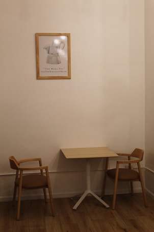 A small, minimalist interior scene features a wooden table with two matching wooden chairs with brown cushions on either side. A framed artwork of a Moka pot hangs on the beige wall above the table, adding a touch of vintage charm. The wooden floor complements the warm, neutral tones of the furniture and wall.