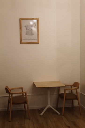 A small, minimalist interior scene features a wooden table with two matching wooden chairs with brown cushions on either side. A framed artwork of a Moka pot hangs on the beige wall above the table, adding a touch of vintage charm. The wooden floor complements the warm, neutral tones of the furniture and wall.