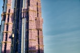 A tall building under construction is wrapped with purple scaffolding covers, with visible supportive structures and sections of glass facades. The sky is clear, enhancing the contrasting colors.