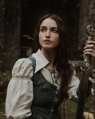 A woman with long, dark hair is dressed in a medieval-style outfit with a white blouse and green vest. She holds an ornate sword with a golden hilt, and her expression is contemplative. The background is filled with foliage and a stone structure, giving a forest-like atmosphere.