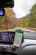 A car dashboard with a mounted smartphone displaying a navigation map is in focus. The car's entertainment system screen shows vehicle settings. In the background, a scenic road with autumn foliage can be seen through the windshield.