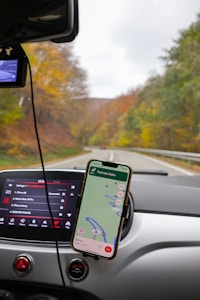 A car dashboard with a mounted smartphone displaying a navigation map is in focus. The car's entertainment system screen shows vehicle settings. In the background, a scenic road with autumn foliage can be seen through the windshield.