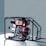 A portable diesel engine generator encased within a black metal frame, featuring a combination of silver, red, and black components. The engine housing displays branding and instructional graphics. The backdrop is a plain, muted gray-blue surface.
