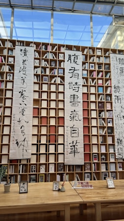 A wooden shelf structure filled with numerous books is adorned with vertical scrolls featuring Chinese calligraphy. The background includes a large window showing a clear blue sky, allowing natural light to illuminate the scene. Below the shelves, there is a long wooden table with various small items and signs placed on it.