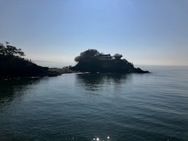 A serene coastal scene with a small island or promontory covered in trees and a traditional building perched above. The sky is clear with the sun casting a gentle light over the calm sea. A group of people appear to be walking along a narrow path on the island.