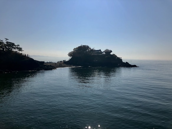 A serene coastal scene with a small island or promontory covered in trees and a traditional building perched above. The sky is clear with the sun casting a gentle light over the calm sea. A group of people appear to be walking along a narrow path on the island.