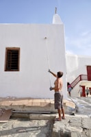 A shirtless man is painting the exterior wall of a white building using a paint roller on a long pole. The building has a wooden window on the left and another entrance with a red railing on the right. There are some cardboard pieces on the floor next to the wall, and a few other people are walking in the background.