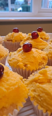 A close-up view of several cupcakes topped with yellow frosting and a single red cherry on top of each. The cupcakes are arranged on a cooling rack in a kitchen setting with a window in the background, showing a blurred view of greenery and a car.
