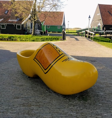 A large, yellow wooden clog sculpture is prominently displayed on a cobblestone path. In the background, there are traditional Dutch houses with distinct red-tiled roofs and wooden walls. The scene is set on a clear day with a few small trees and a green grassy area visible.