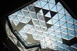 A geometric glass ceiling composed of triangular panes, allowing sunlight to stream through. The view leads upwards, displaying part of a tall building through the transparent sections. Shadows are cast on the frame, adding a dynamic element to the scene.