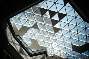 A geometric glass ceiling composed of triangular panes, allowing sunlight to stream through. The view leads upwards, displaying part of a tall building through the transparent sections. Shadows are cast on the frame, adding a dynamic element to the scene.