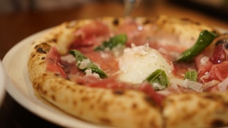 A close-up shot of a pizza topped with creamy cheese, vibrant green vegetables, and thin slices of cured meat. The crust appears golden and lightly charred.