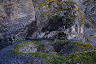 A narrow, rocky mountain path curves around a steep cliff face covered in moss and grass. A hiker wearing dark clothing with a backpack and trekking poles stands on the path, gazing at the impressive rock formations. The surrounding environment is rugged and textured, with layers of rock and patches of greenery.
