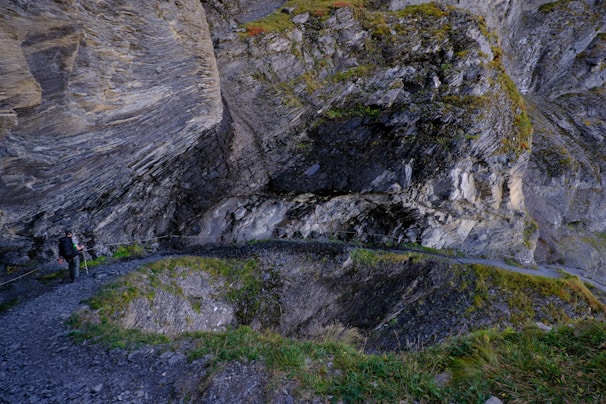 A narrow, rocky mountain path curves around a steep cliff face covered in moss and grass. A hiker wearing dark clothing with a backpack and trekking poles stands on the path, gazing at the impressive rock formations. The surrounding environment is rugged and textured, with layers of rock and patches of greenery.