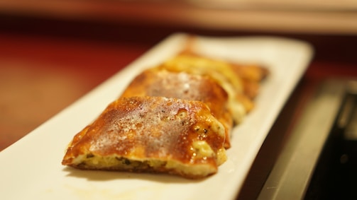 A close-up view of several pieces of golden-brown baked pastry items arranged on a white rectangular plate. The pastries appear to be filled with a savory or cheesy filling, and the crust looks crispy and well-cooked.