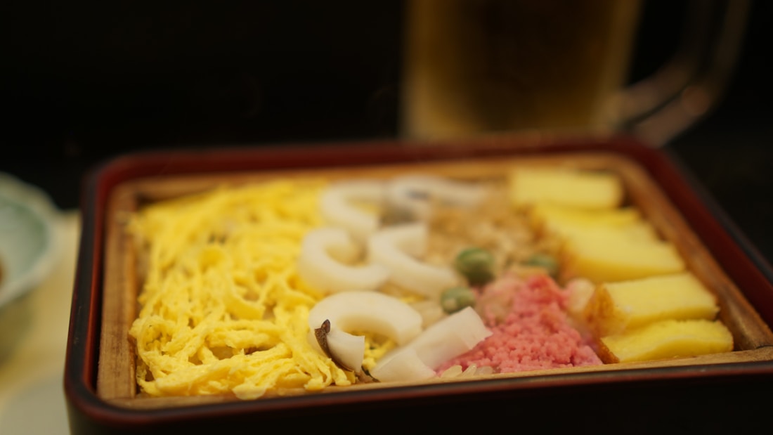 A bento box features a variety of foods, including shredded egg, white seafood slices, green peas, diced pink pickles, and yellow tamagoyaki arranged neatly in sections. The background is slightly blurred, with a beverage glass partially visible.