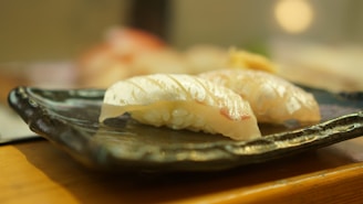 Two pieces of sushi placed on a dark, rectangular ceramic plate with a shiny finish. The sushi is topped with translucent, white fish slices over a mound of rice. The setting has a warm, natural light, creating a peaceful and inviting atmosphere.