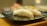 Two pieces of sushi placed on a dark, rectangular ceramic plate with a shiny finish. The sushi is topped with translucent, white fish slices over a mound of rice. The setting has a warm, natural light, creating a peaceful and inviting atmosphere.