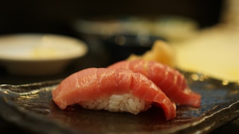 Two pieces of sushi with raw fish slices on a bed of rice are placed on a textured, dark ceramic plate. The background is slightly out of focus, featuring round white and dark bowls.