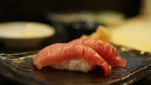 Two pieces of sushi with raw fish slices on a bed of rice are placed on a textured, dark ceramic plate. The background is slightly out of focus, featuring round white and dark bowls.