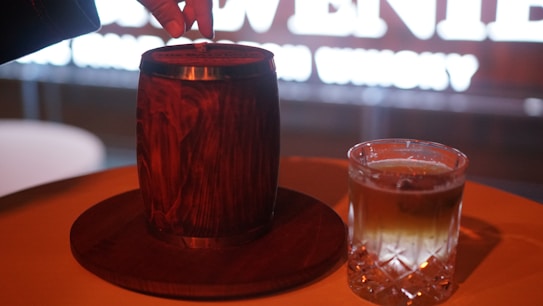 A wooden barrel placed on a round wooden tray with a hand reaching toward its top. Next to it, a glass filled with a light brown liquid, likely whiskey, sits on the same surface. The background features out-of-focus bright text.