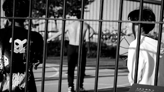 A black and white photograph of a basketball court with three individuals. The scene is viewed through a metal fence, focusing on two people in the foreground. One person holds a basketball while smiling, and another faces away from the camera wearing a shirt with graphic designs. In the background, a shirtless person is captured in motion.