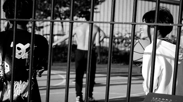 A black and white photograph of a basketball court with three individuals. The scene is viewed through a metal fence, focusing on two people in the foreground. One person holds a basketball while smiling, and another faces away from the camera wearing a shirt with graphic designs. In the background, a shirtless person is captured in motion.
