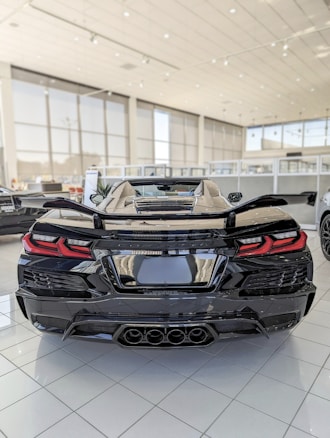 A sleek black sports car is prominently displayed in a spacious showroom. The car is highly polished, with a glossy exterior and stylish rear design featuring distinctive taillights and quad exhaust pipes. The showroom is well-lit with large windows and tiled flooring, creating an ambience of luxury and modernity.