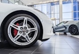 A close-up of the front wheel of a white sports car is in the foreground, showing detailed alloy rims and a red brake caliper. In the background, there is another grey sports car parked inside a modern showroom with large glass windows and a tiled floor.