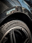 Close-up of a car tire with the brand 'Michelin' and model 'Pilot Sport' clearly visible on the rubber. The rim is sleek and shiny, surrounded by a metallic car body that has some visible water droplets. The background shows a partial reflection on the car's surface.