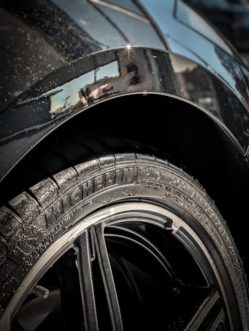 Close-up of a car tire with the brand 'Michelin' and model 'Pilot Sport' clearly visible on the rubber. The rim is sleek and shiny, surrounded by a metallic car body that has some visible water droplets. The background shows a partial reflection on the car's surface.