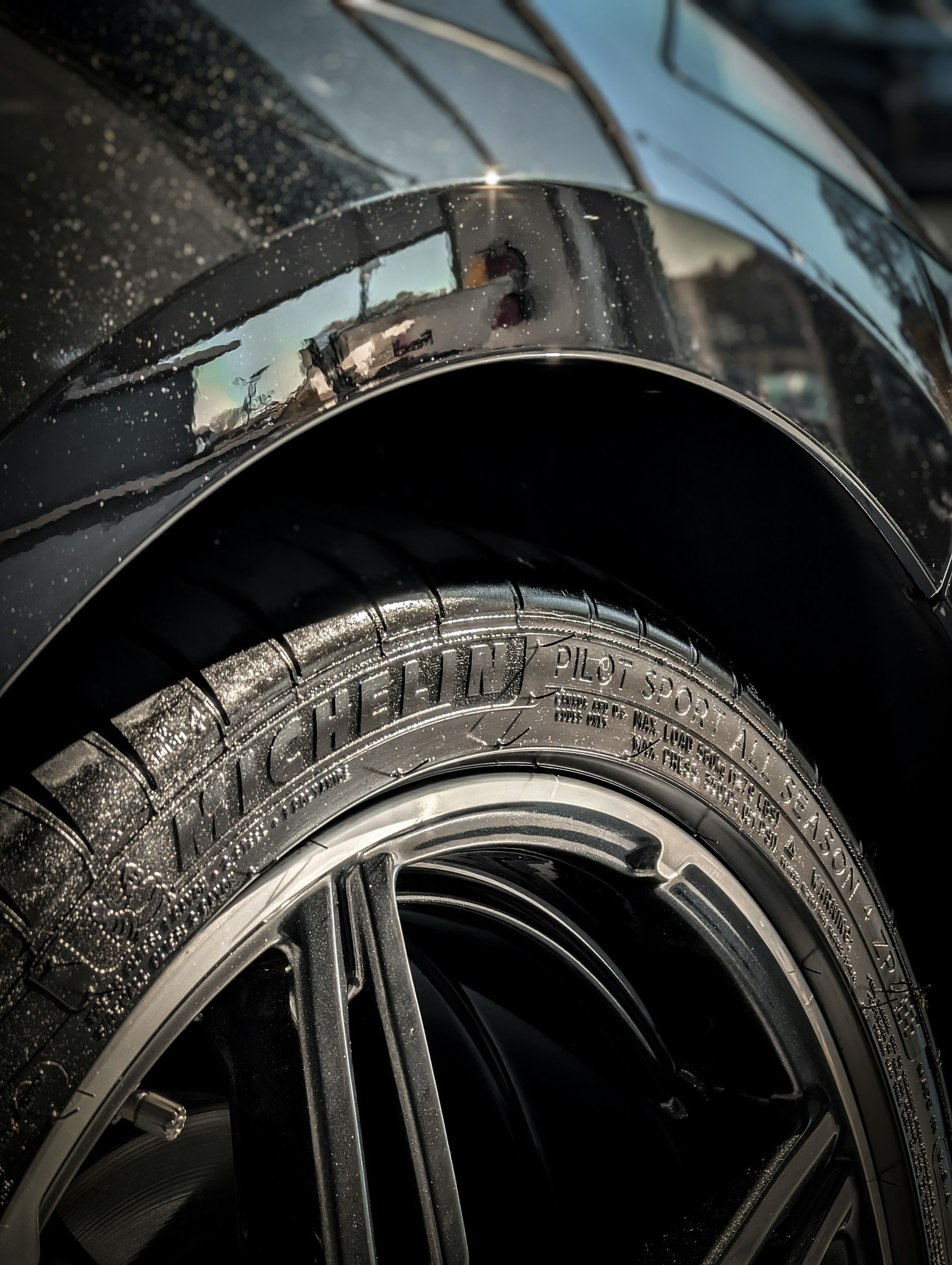 Close-up of a car tire with the brand 'Michelin' and model 'Pilot Sport' clearly visible on the rubber. The rim is sleek and shiny, surrounded by a metallic car body that has some visible water droplets. The background shows a partial reflection on the car's surface.