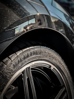 Close-up of a car tire with the brand 'Michelin' and model 'Pilot Sport' clearly visible on the rubber. The rim is sleek and shiny, surrounded by a metallic car body that has some visible water droplets. The background shows a partial reflection on the car's surface.