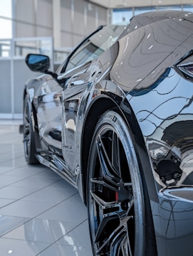 A close-up view of a sleek, modern sports car in a showroom, focusing on the rear wheel with its shiny, intricate rim detailing. The reflective surface of the car highlights the luxurious and polished nature of the vehicle, with glimpses of the showroom environment subtly visible in the car’s bodywork.