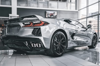 A sleek silver sports car displayed in a modern showroom with large windows. The vehicle features distinctively designed tail lights and black, alloy wheels. The floor of the showroom is polished, reflecting the car's streamlined body.